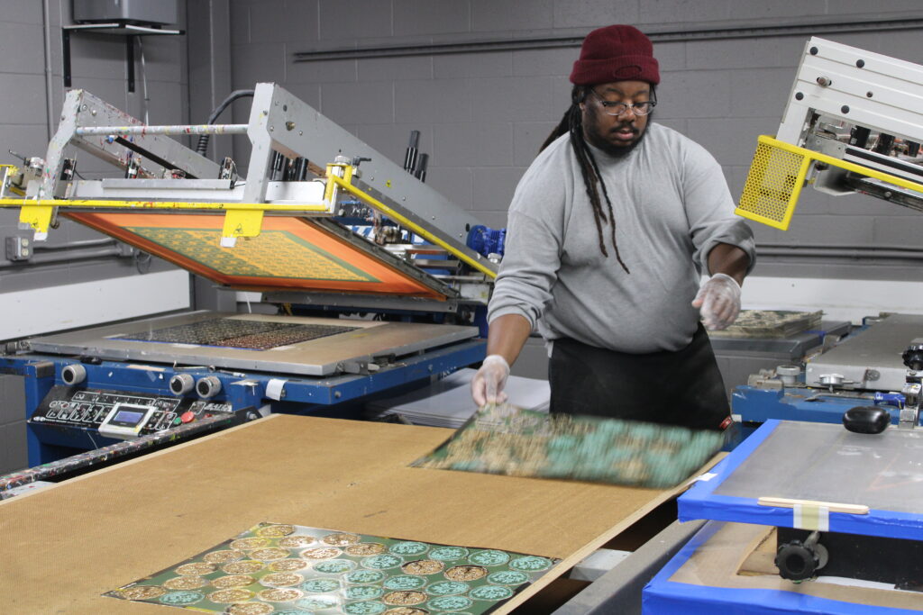 An employee pursuing a career at ChemArt carefully handles a printed sheet on a production table in our manufacturing facility, working with screen printing equipment.