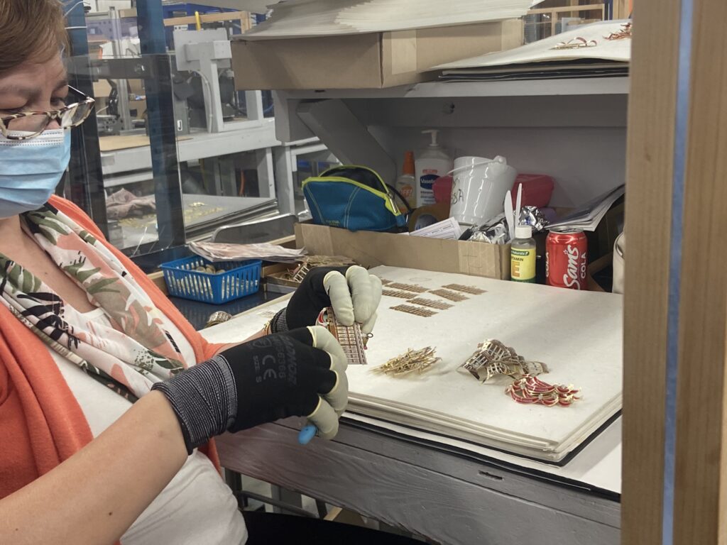 An employee wearing gloves and a mask carefully assembles or inspects intricate brass ornaments at a workbench filled with tools and supplies.